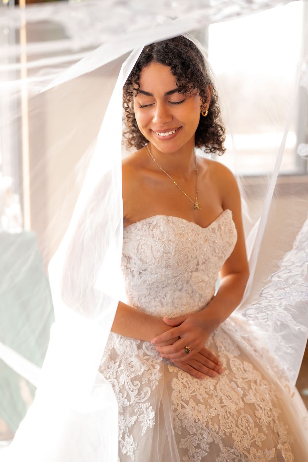 Beautiful bride in white lace strapless wedding dress and veil smiles with eyes closed in sunlit setting.