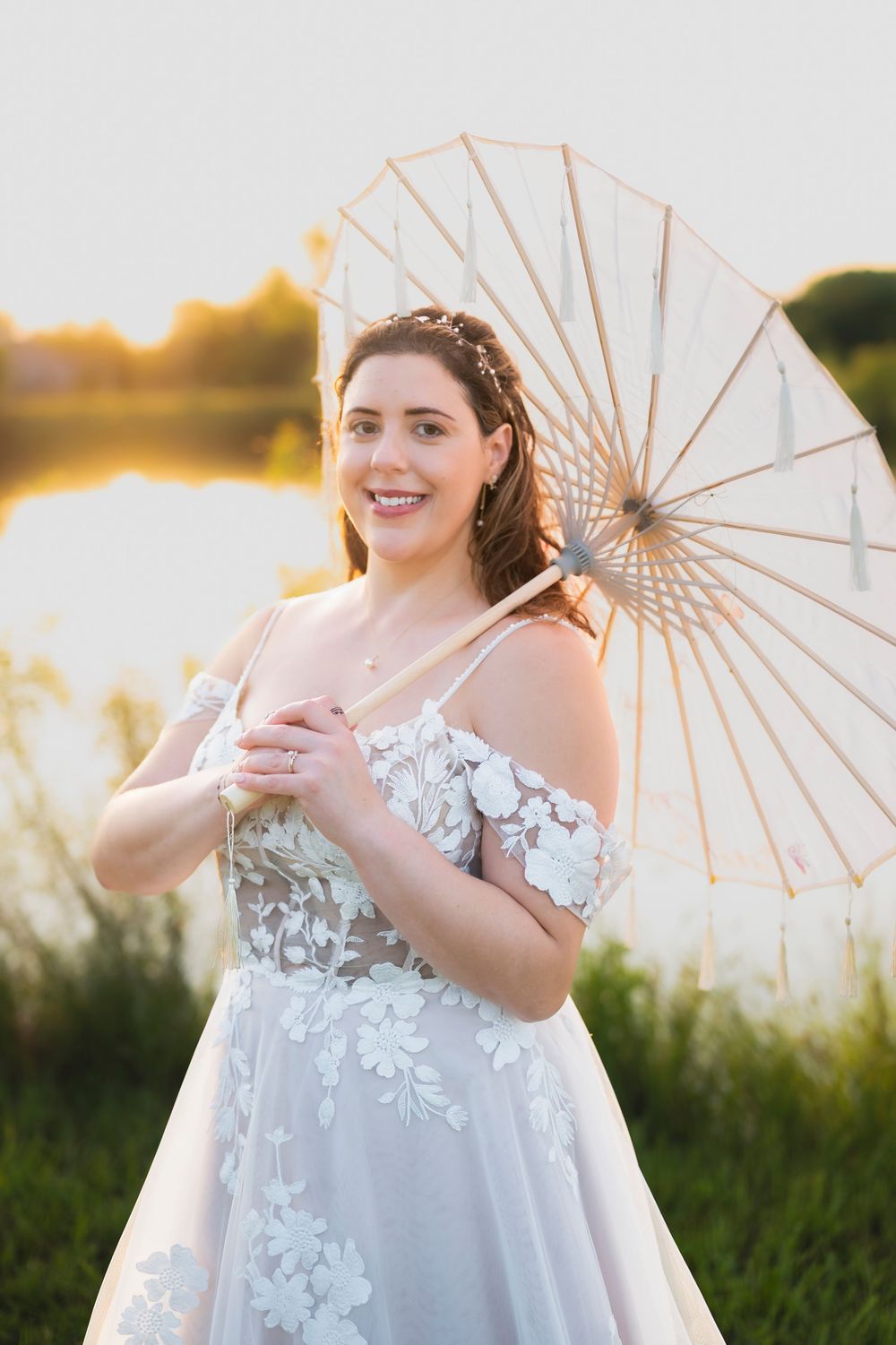 A bride poses outdoors at sunset in a white floral lace dress with an umbrella parasol in a summer setting.