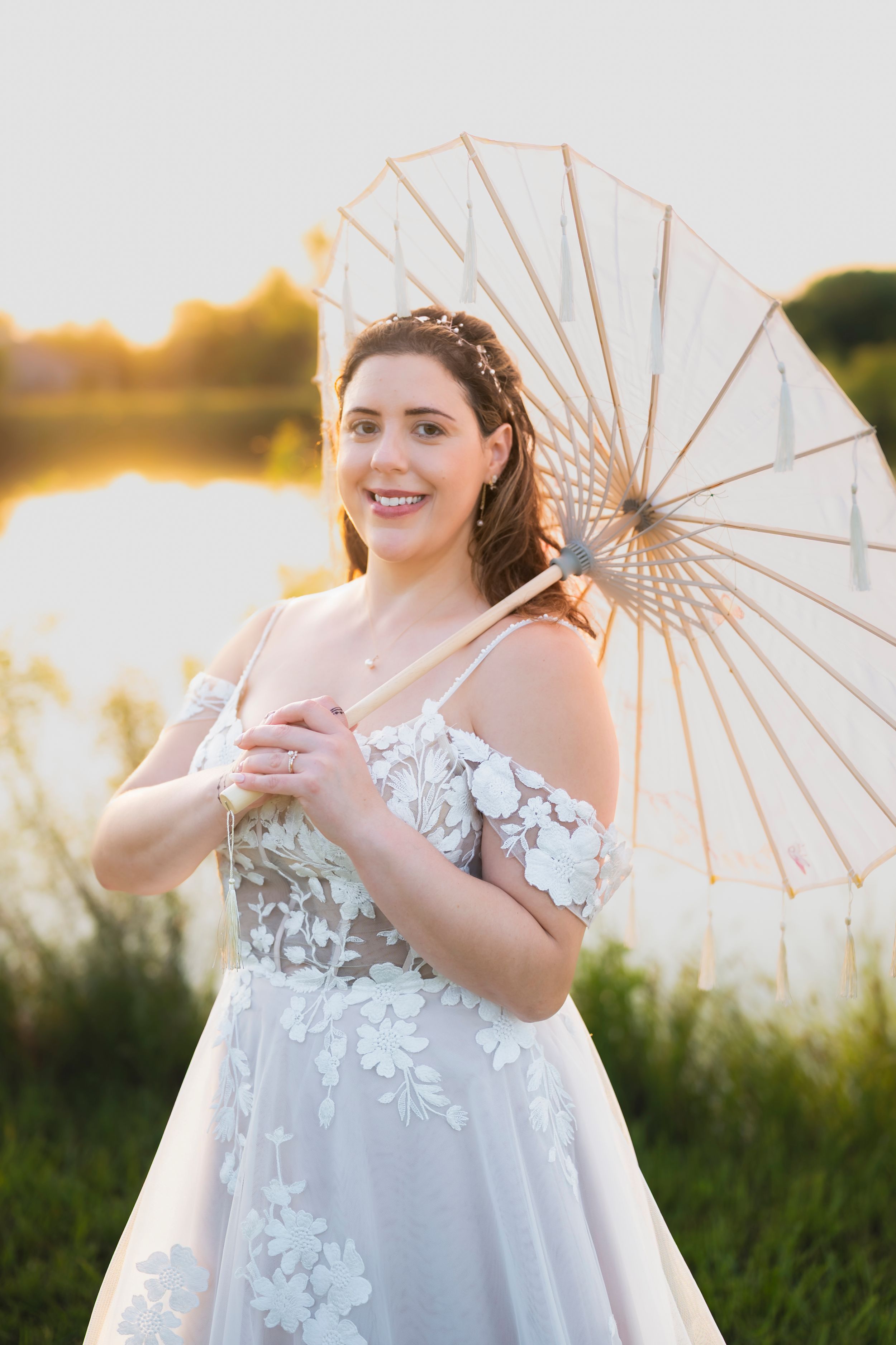 A bride poses outdoors at sunset in a white floral lace dress with an umbrella parasol in a summer setting.