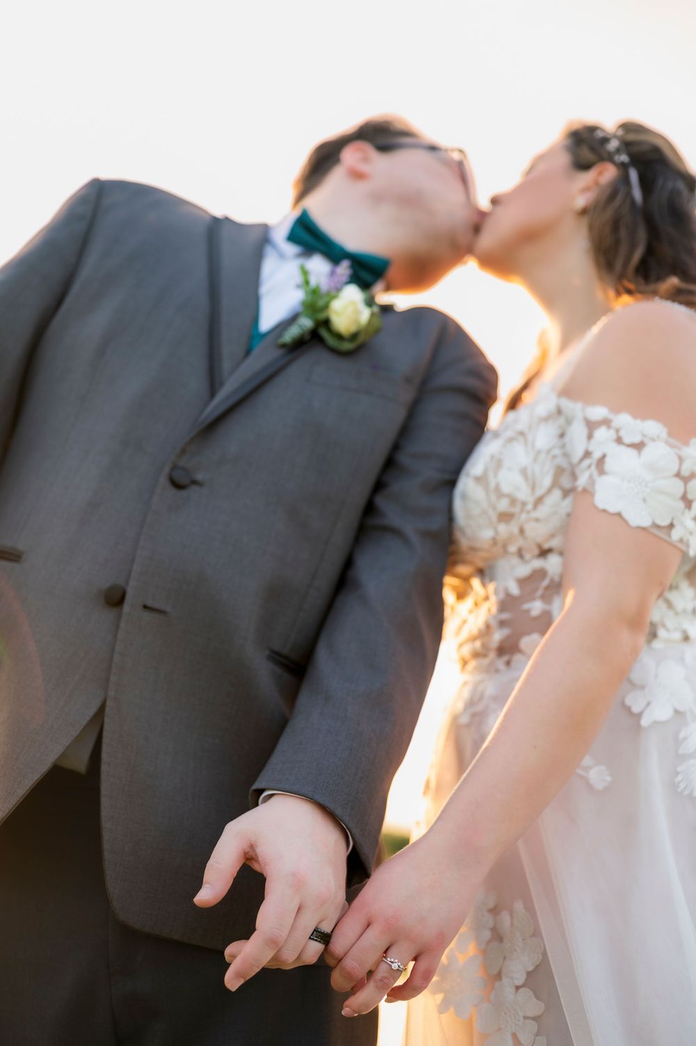 Close-up wedding photo details showing hands with rings against formal attire in warm lighting.
