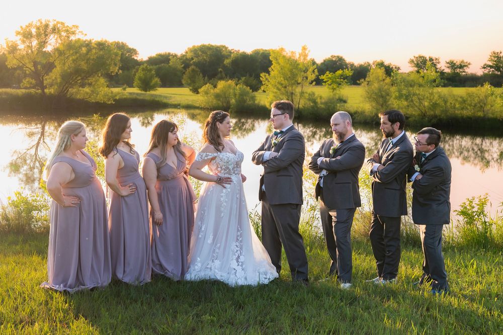 Wedding party sitting together on grassy lawn at sunset with bridesmaids in blush dresses.