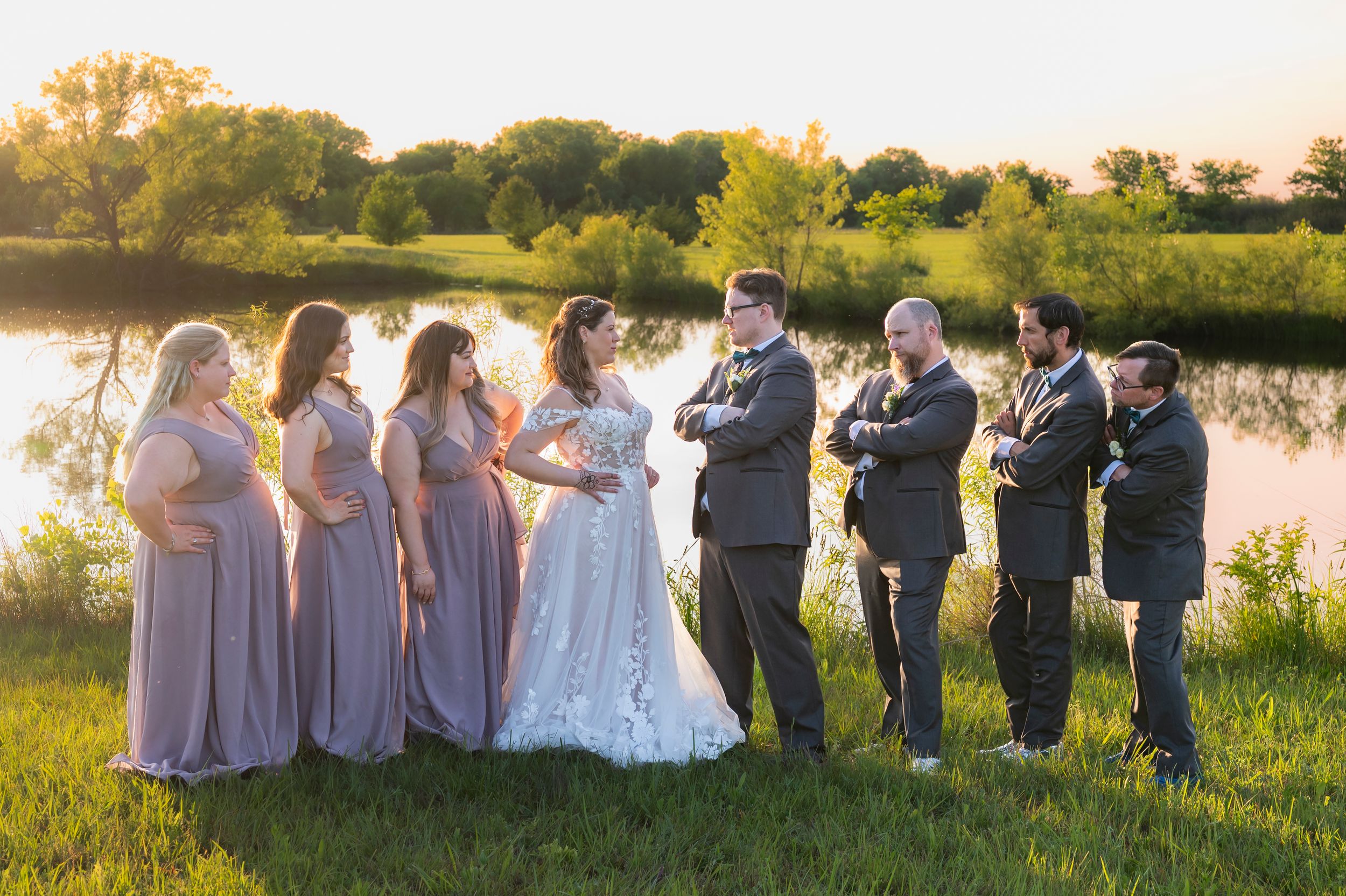 Wedding party sitting together on grassy lawn at sunset with bridesmaids in blush dresses.