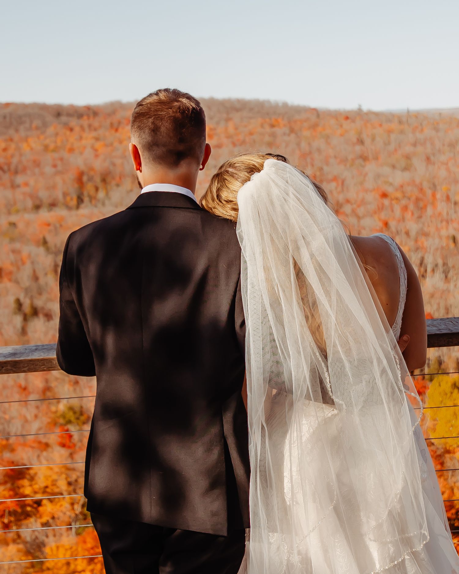 A couple in wedding attire looks out over an autumn landscape from a viewpoint.