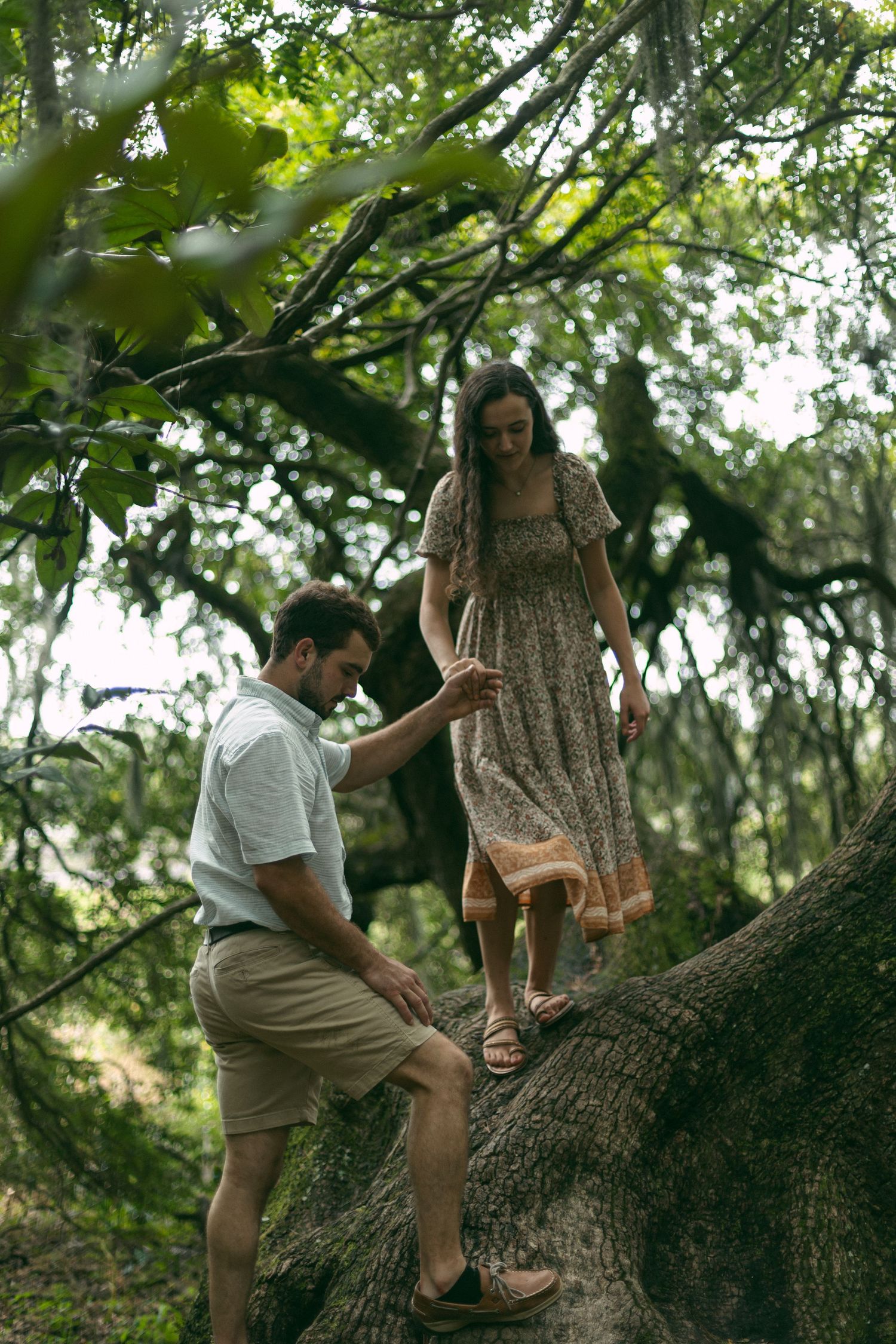 A couple carefully navigates their way on a large tree trunk in a lush garden setting.