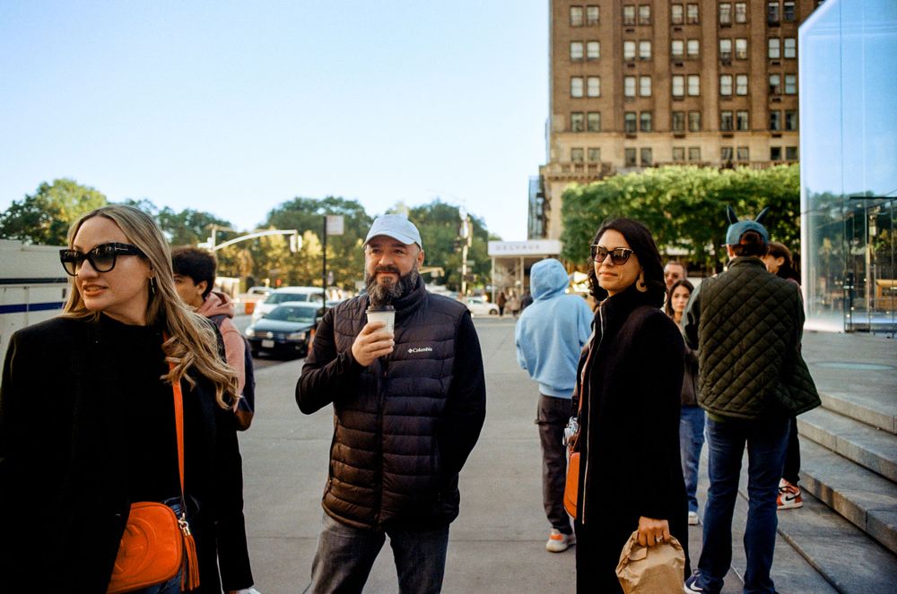 People gather on a sidewalk in an urban setting on a sunny day.
