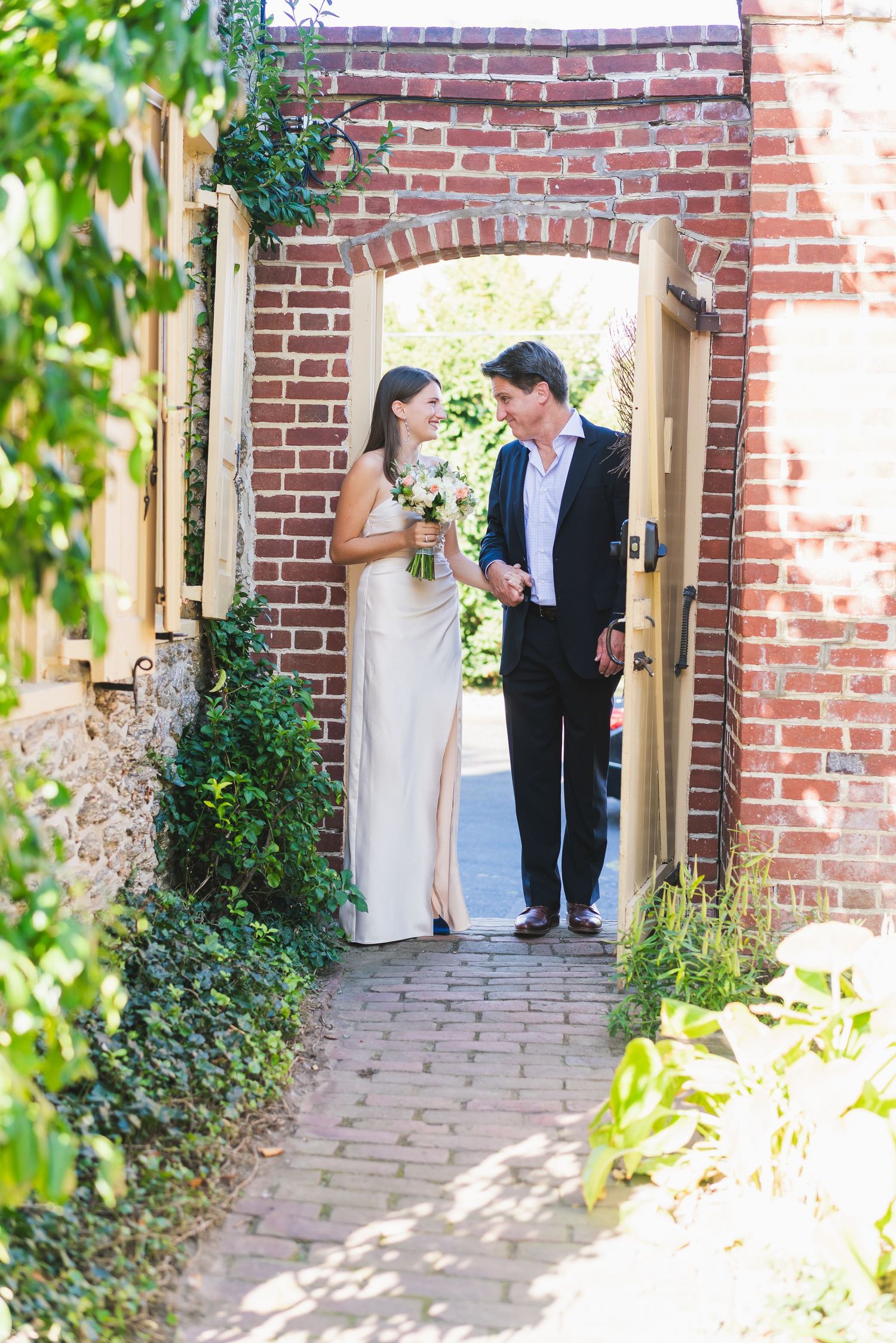 A couple in formal attire stands in a romantic brick archway surrounded by greenery.
