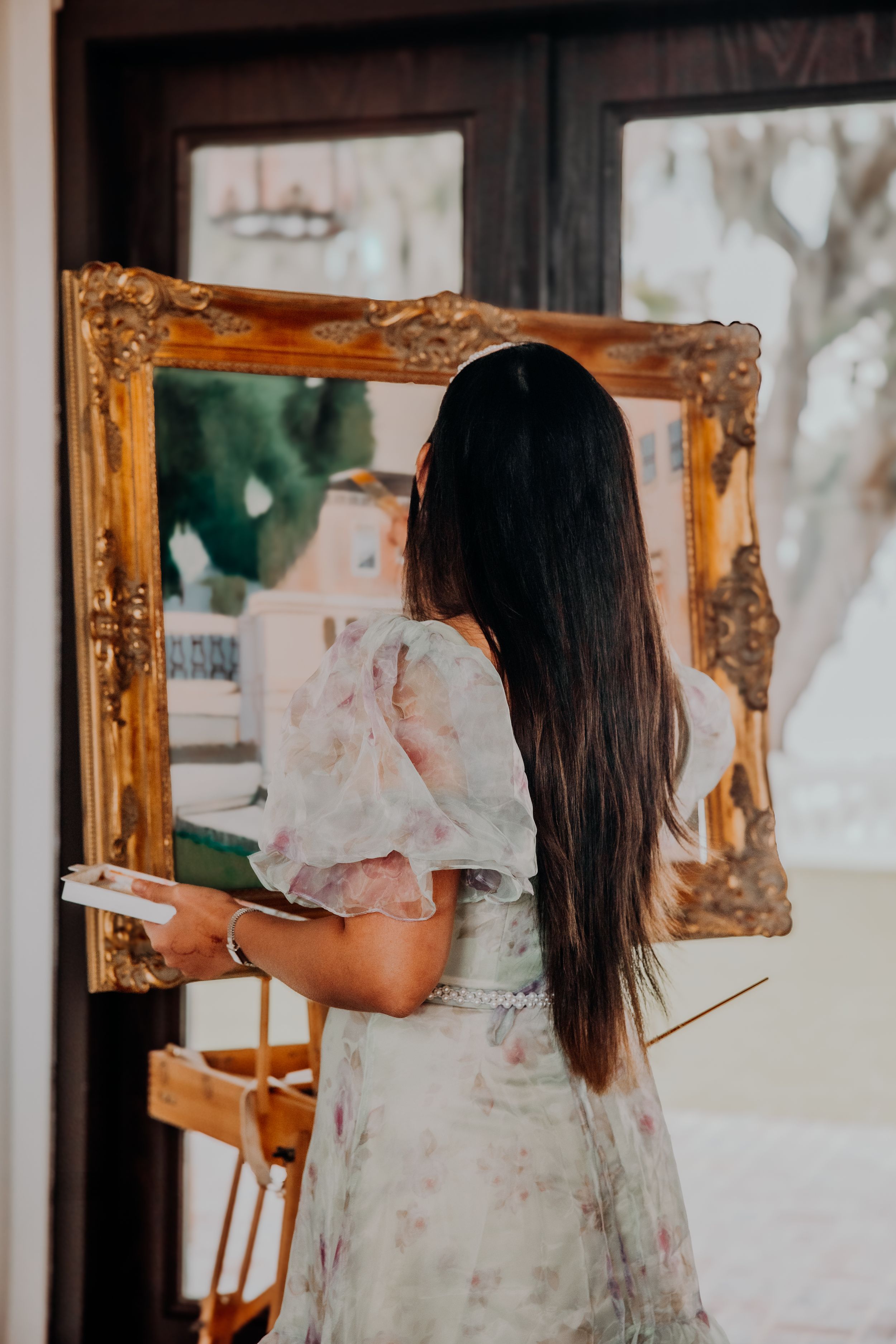 A person in a vintage dress is reflected in multiple ornate gold mirrors in a bright room.