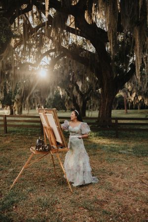 A painter in a floral dress creates artwork under moss-covered oak trees during golden hour.