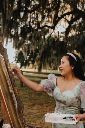 An artist in a floral dress paints enthusiastically at an outdoor easel under oak trees.