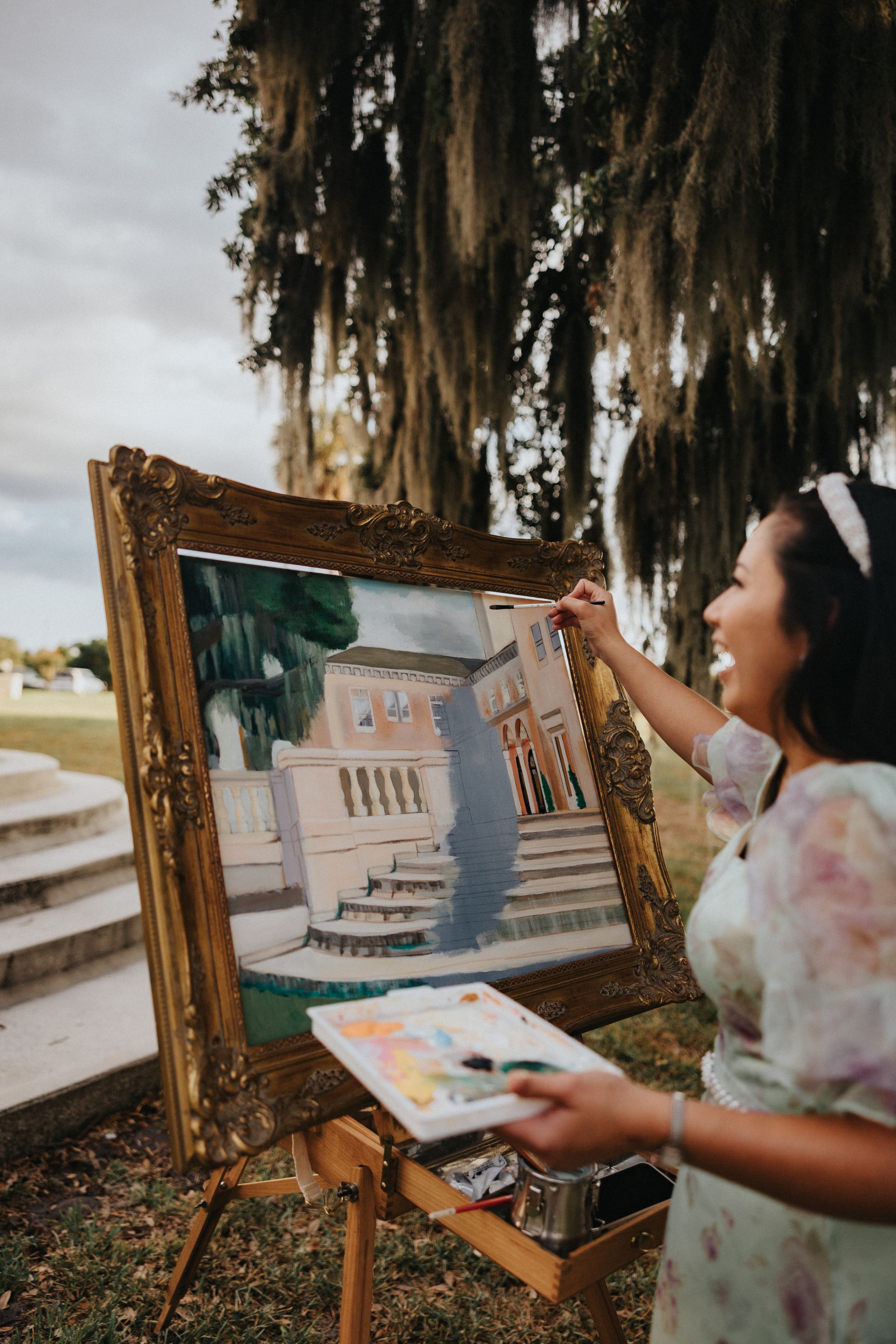 Artist painting a scenic building landscape on an easel outdoors under Spanish moss draped trees.