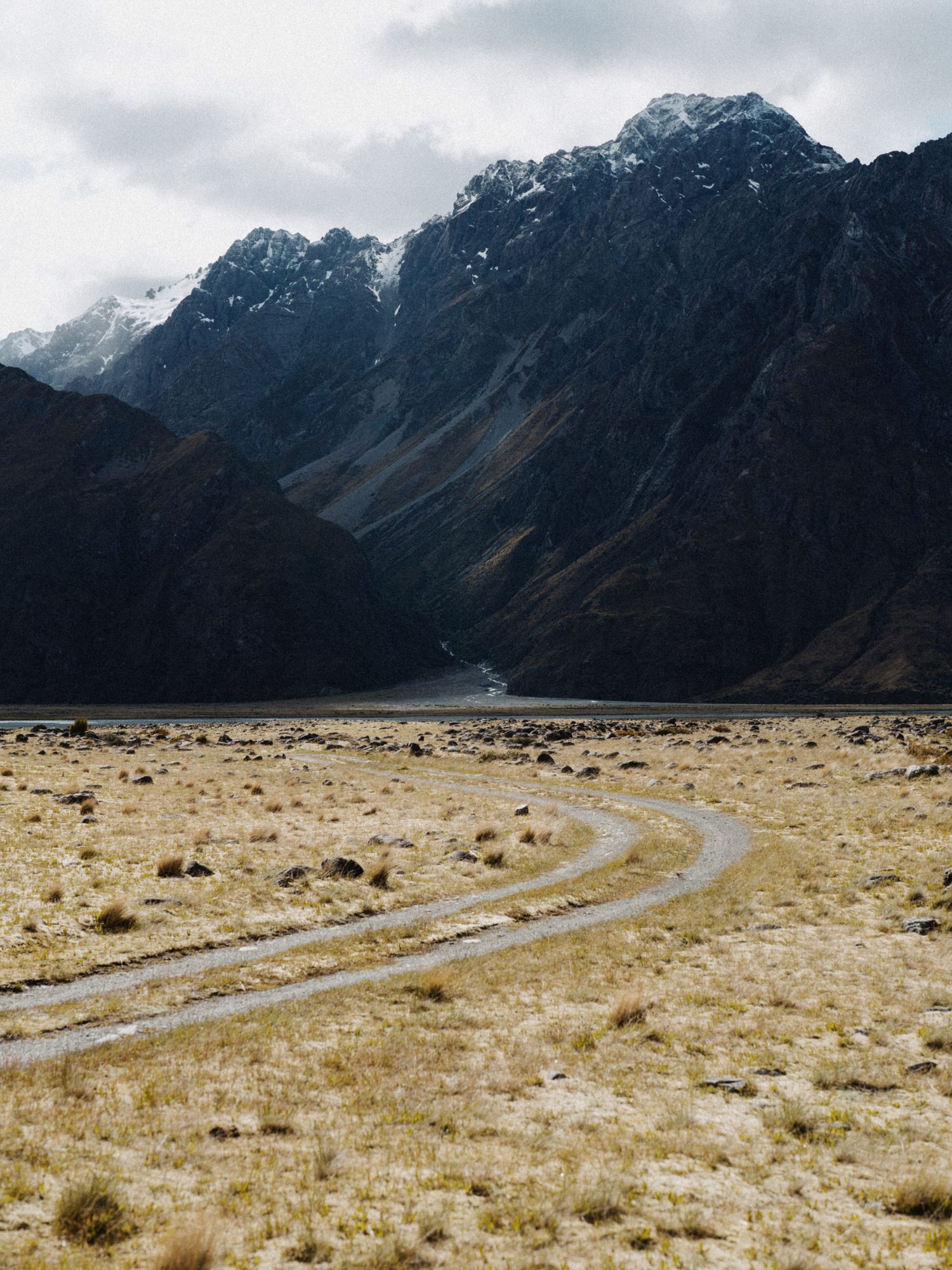 A dirt road curves through arid grassland towards dramatic mountain peaks.