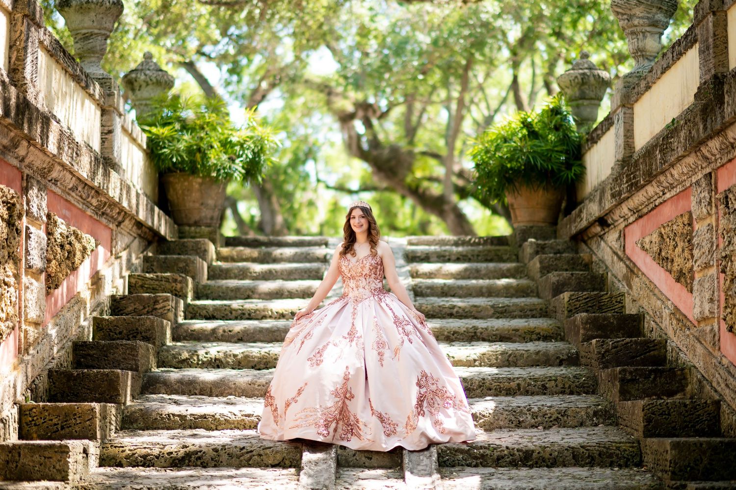 A pink floral ballgown flows gracefully on ornate garden steps surrounded by lush greenery and stone architecture.