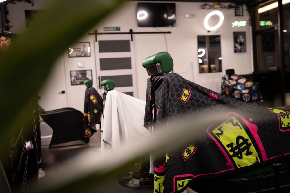 Barbershop chairs covered with vibrant patterned capes viewed through green plant leaves.