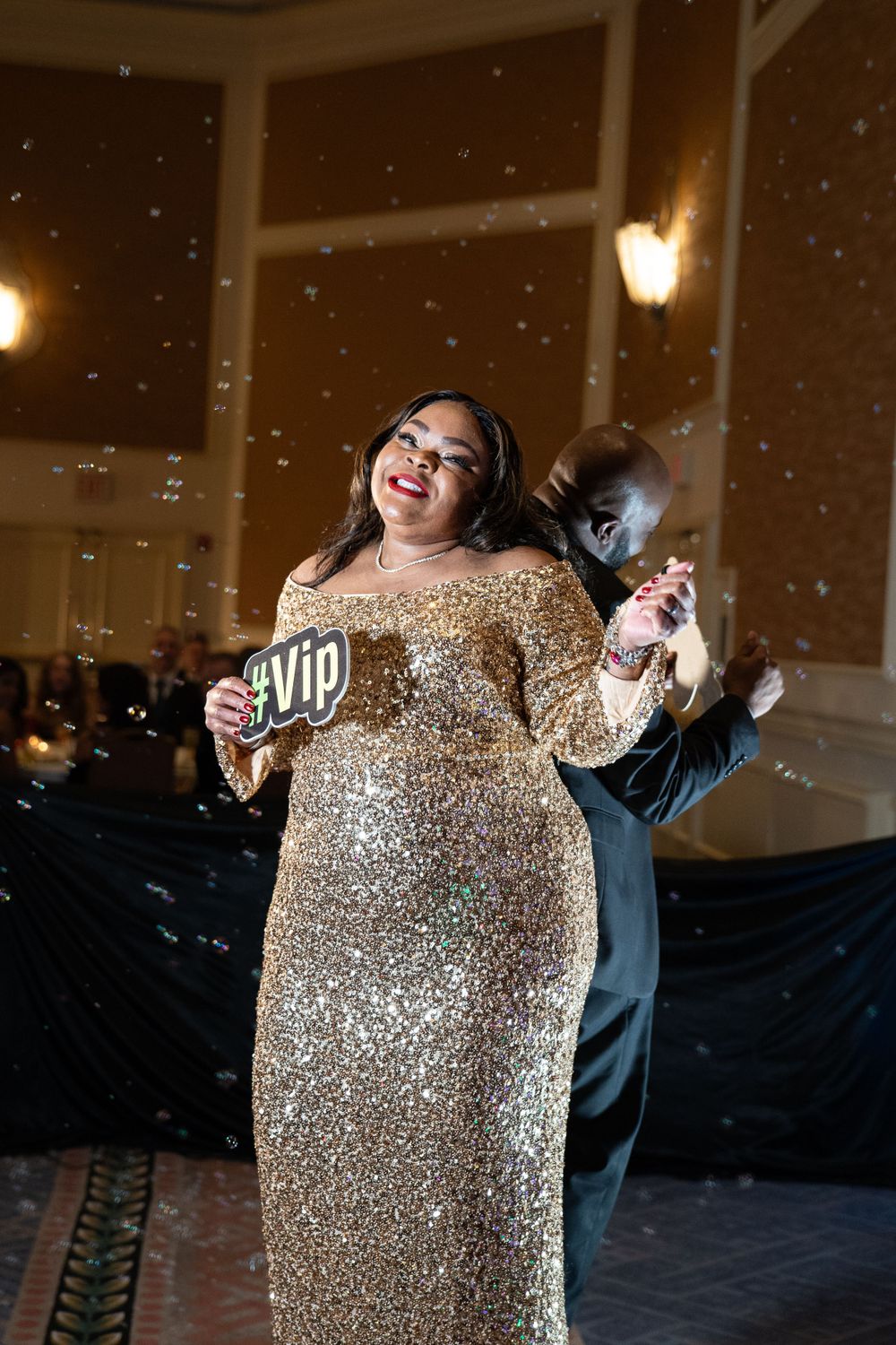 Woman in gold sequin off-shoulder gown holds VIP sign while posing with confetti at gala event.