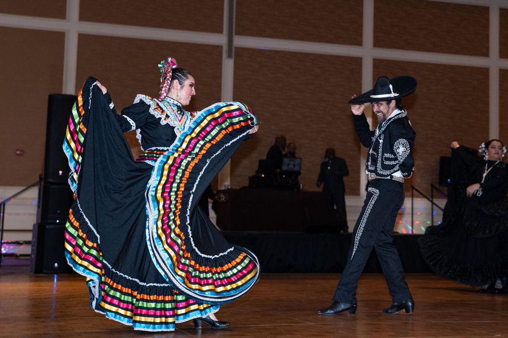 Mexican folklórico dancers perform in vibrant black and multicolored dress with swirling skirt in ballroom.