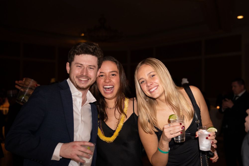 Three smiling young adults pose together holding drinks at a dimly lit evening social event.
