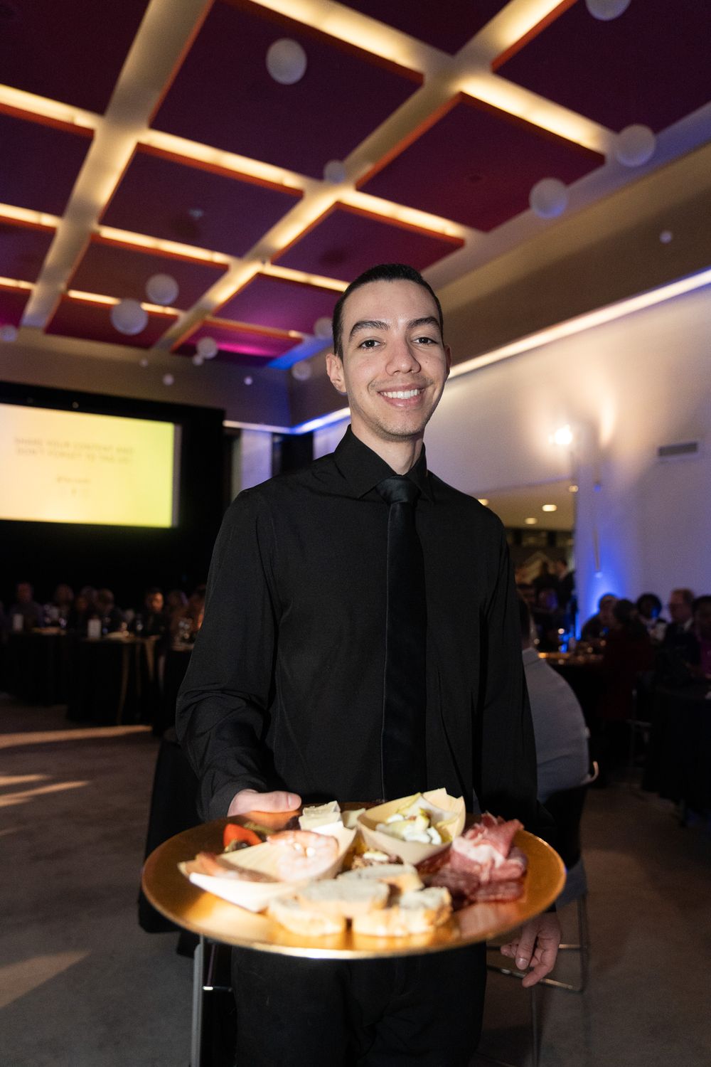 Smiling waiter in black uniform holding a gold tray of assorted charcuterie and cheese appetizers at gala.