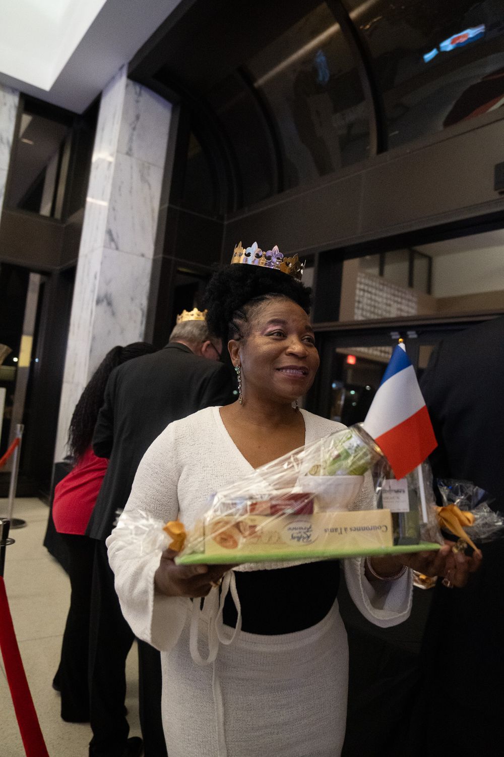 Smiling woman wearing a gold crown holds a gift basket with a French flag at a formal event.
