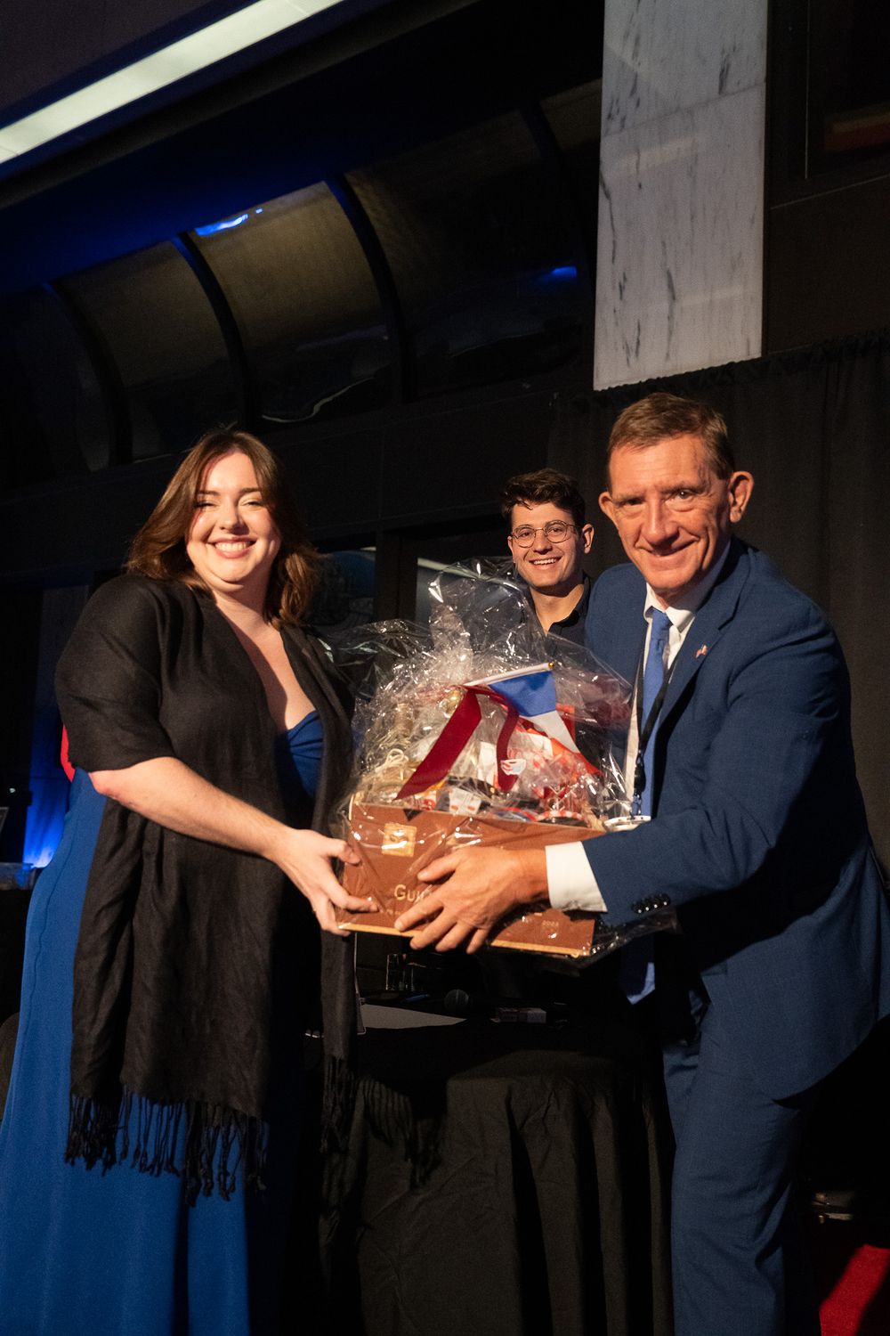 Three people smiling while holding a large gift basket together at a formal evening event.