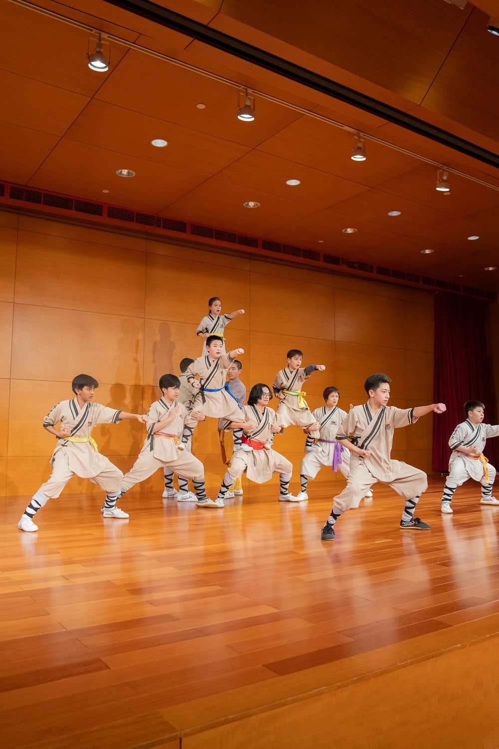 Children perform traditional Chinese martial arts on a wooden stage in matching white uniforms with colorful belts.