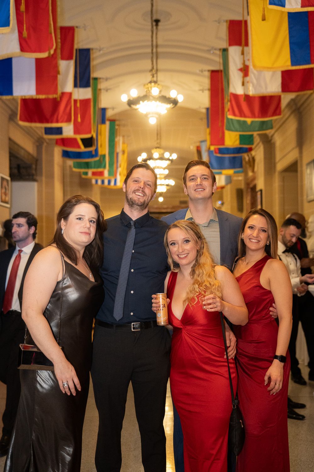 Group of five smiling friends posing in formal attire under international flags in elegant hallway.