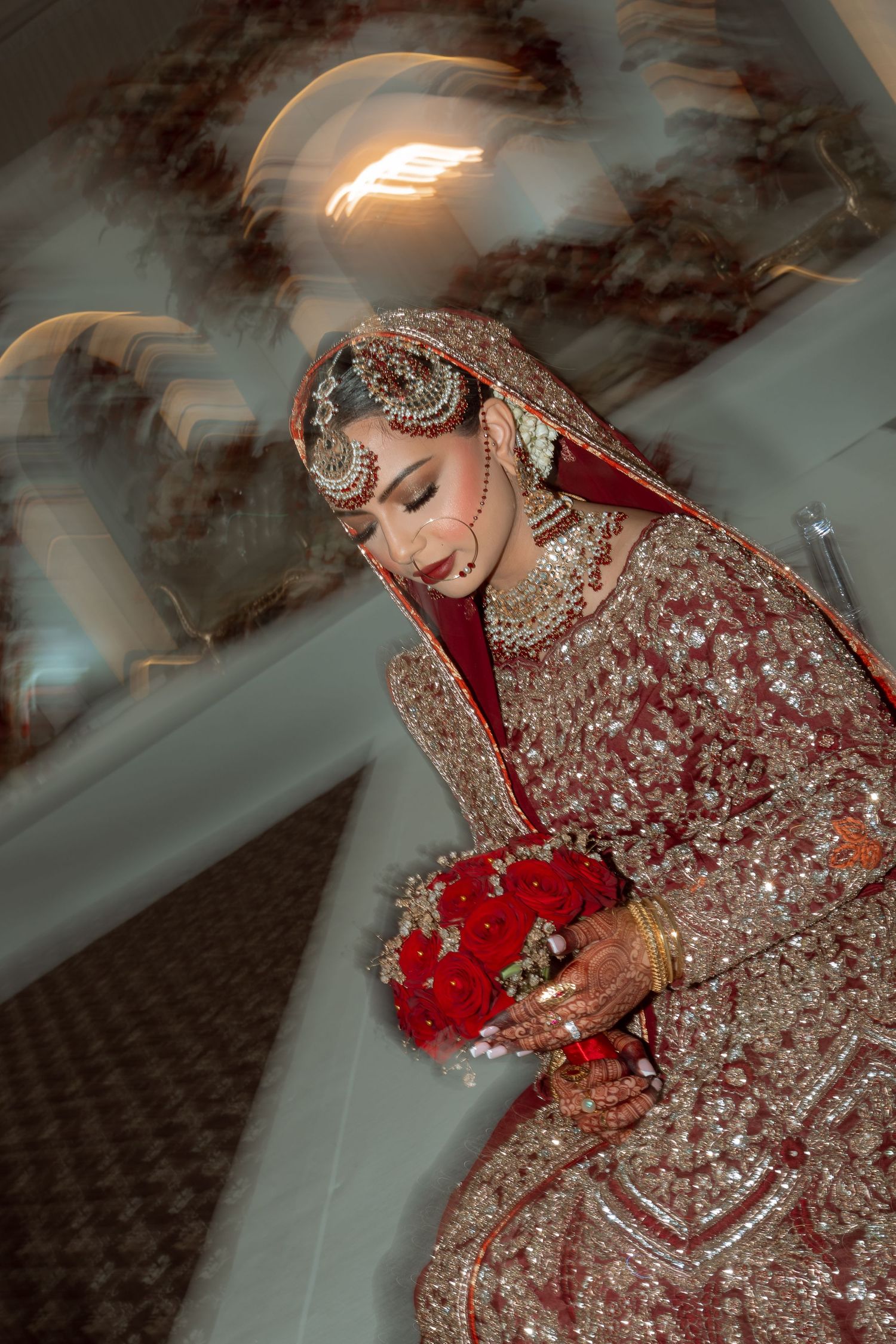 Hands adorned with intricate henna designs and bangles holding a black perfume bottle against a gray background.