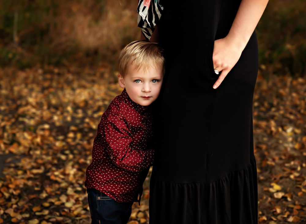 Little blonde boy with blue eyes looking at the camera while staying close to his mom near Airdrie, Alberta