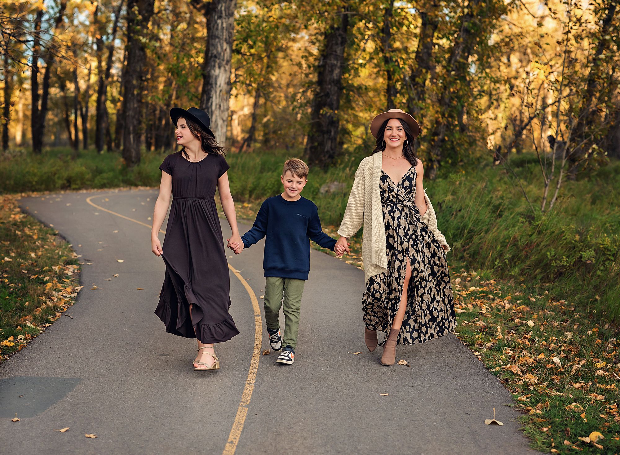 Mom holding her sons hand who is holding his sisters hand walking on a path in Nose Creek Park, Alberta