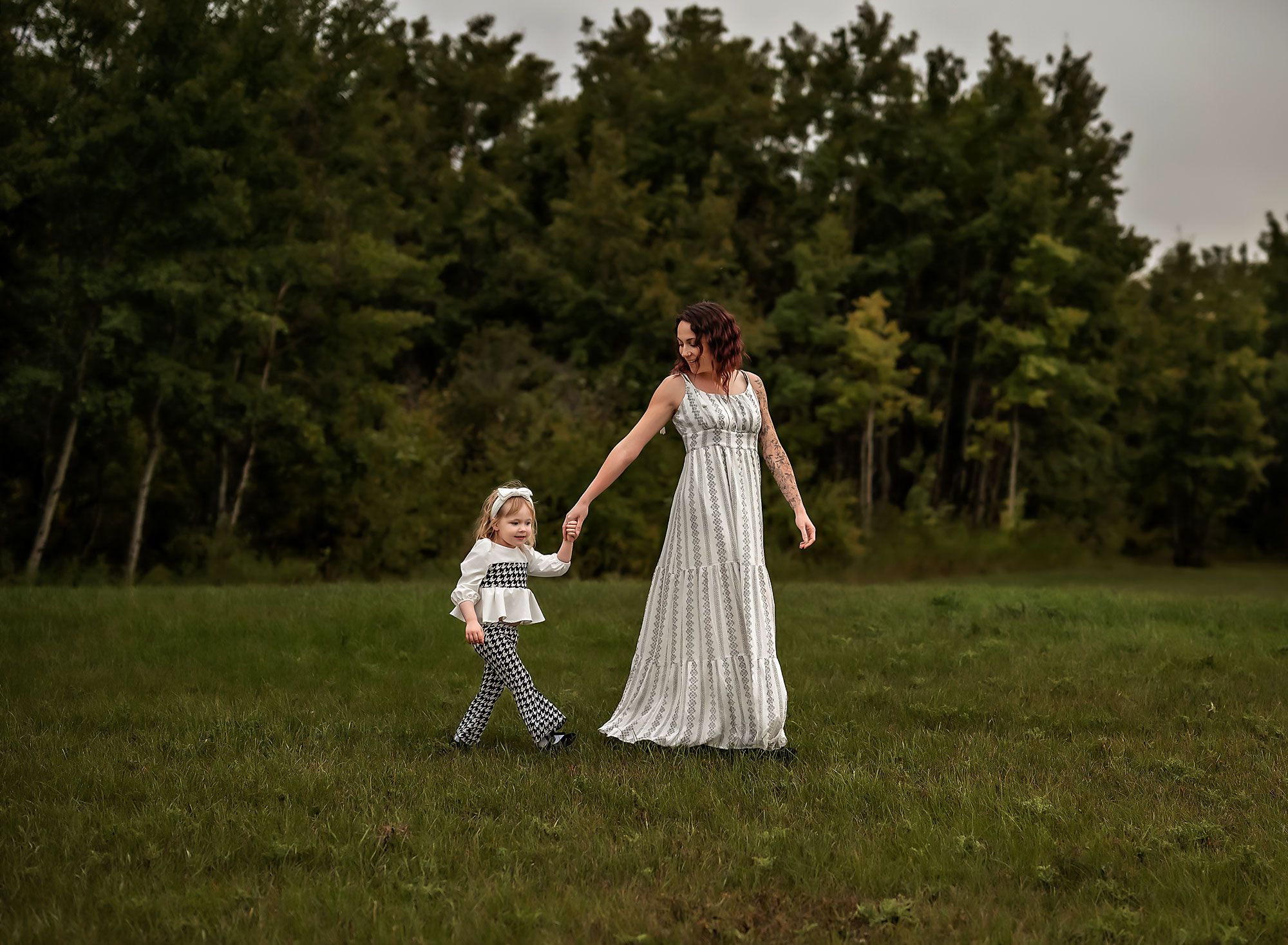 Woman in black and white long dress holding her toddler's hand walking in the forest in Airdrie, Alberta