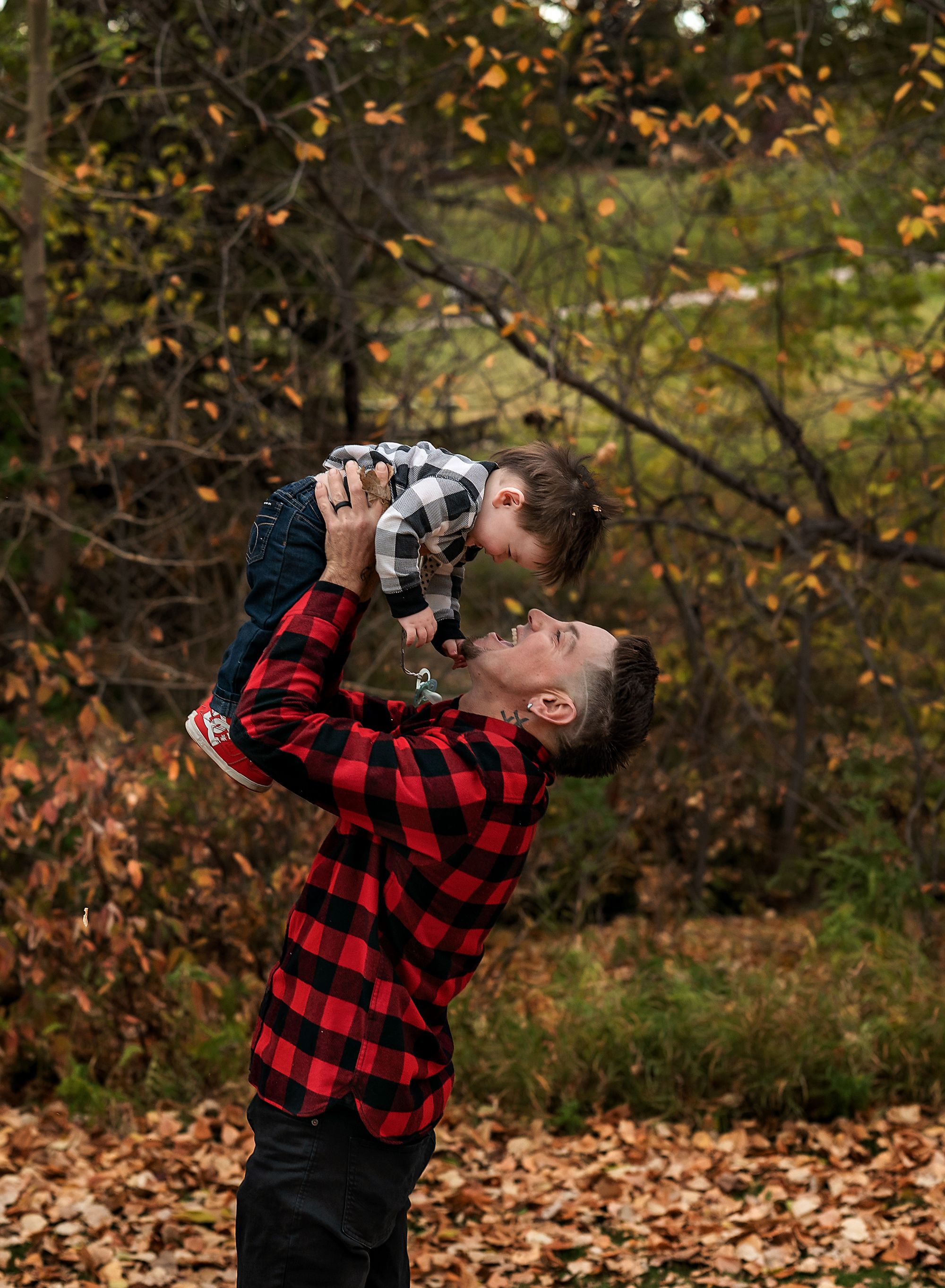 Dad wearing black and white plaid holding his baby boy up in the air looking in his eyes at Confederation Park, Calgary, Alberta