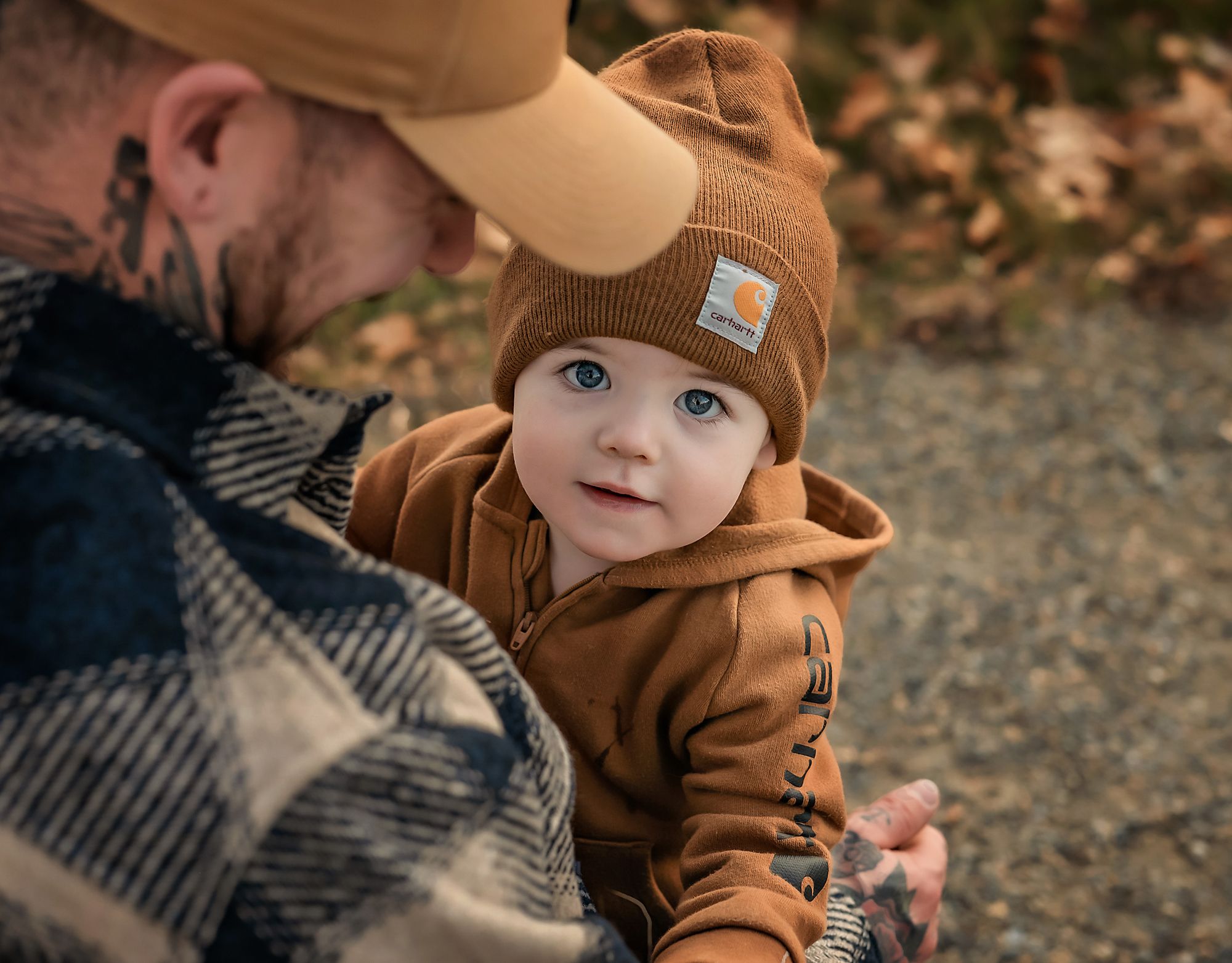 A dad holding his baby boy who is wearing brown carhartt jumpsuit and matching toque in Fish Creek Park, Calgary, Alberta