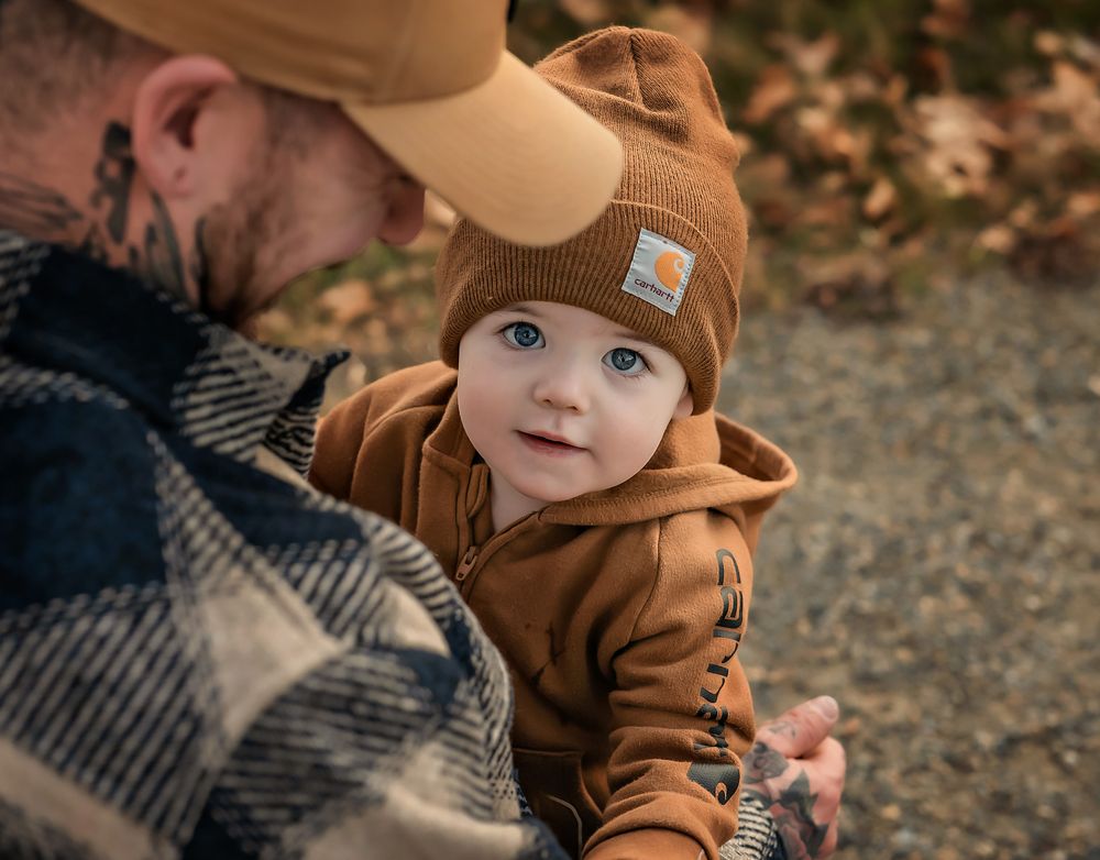 A dad holding his baby boy who is wearing brown carhartt jumpsuit and matching toque in Fish Creek Park, Calgary, Alberta