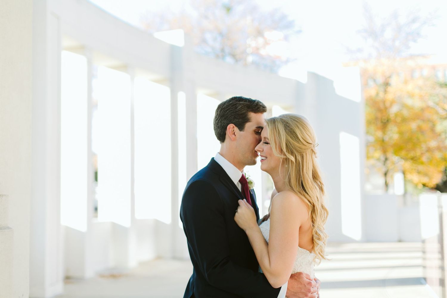 Elegant couple embracing in formal wedding attire against white colonnade architecture.