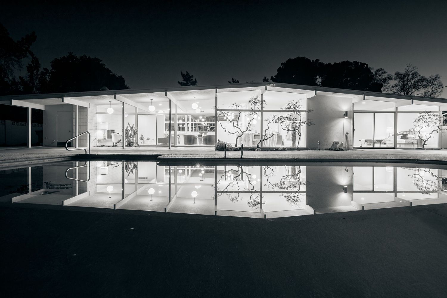 Modern glass-walled pavilion with white exterior illuminated at night, reflecting perfectly in still water.