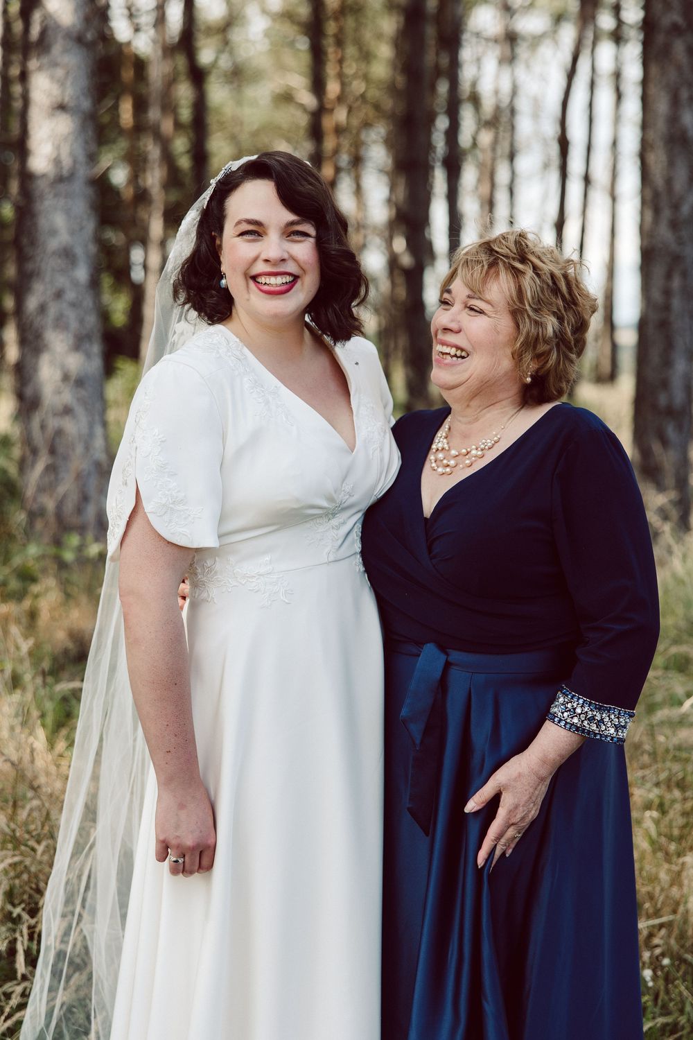 Two women in formal dresses share a joyful moment together in a wooded outdoor setting.