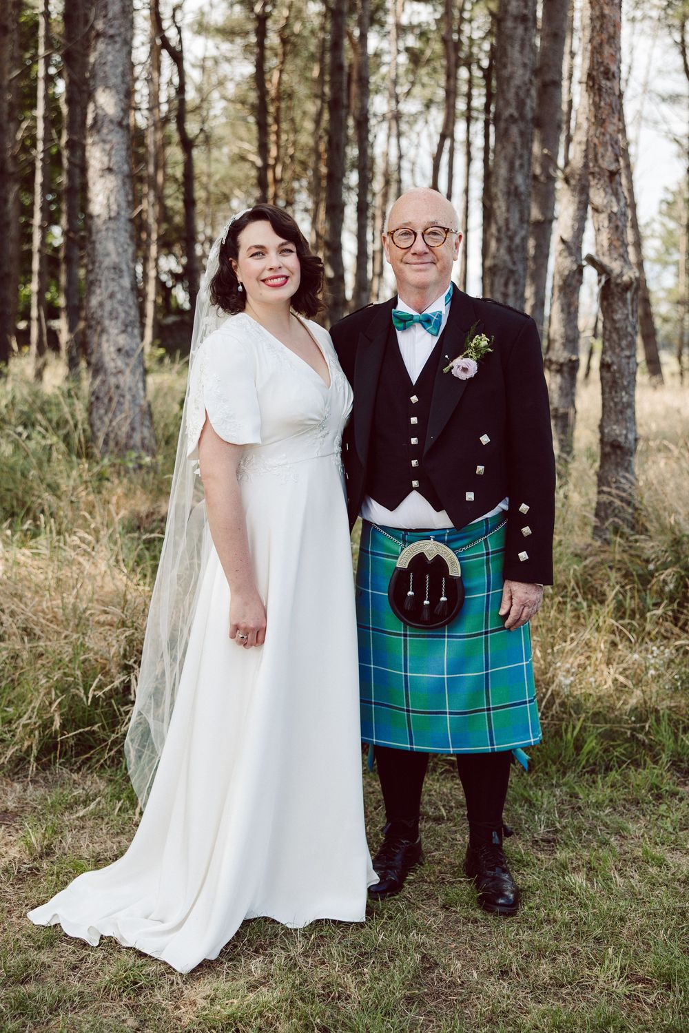 A wedding couple poses in a forest, with the groom wearing traditional Scottish kilt attire in green tartan.