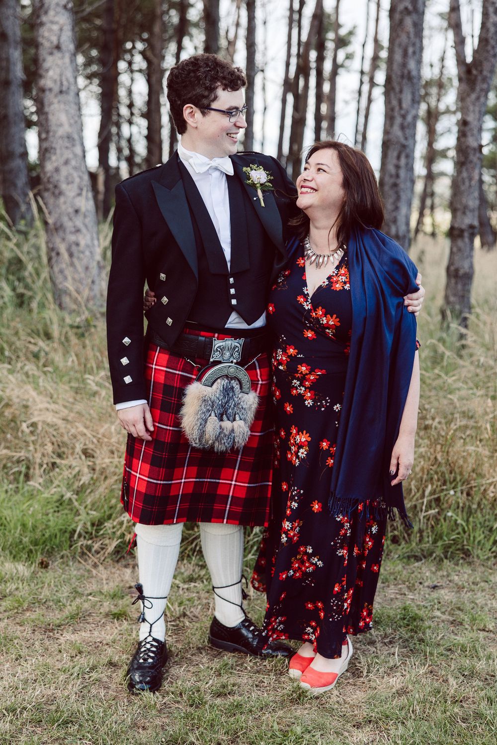 Couple in traditional Scottish attire and formal wear embrace in a wooded setting with red plaid kilt details.