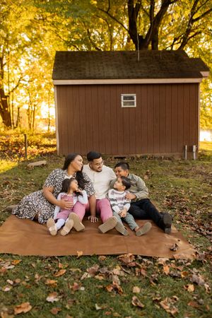 Family lying together on a blanket during golden hour in autumn with fallen leaves and a brown shed in background in Boyds Maryland
