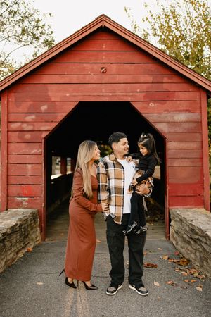 A couple stands together in front of a rustic red covered bridge on an autumn day In Frederick Maryland