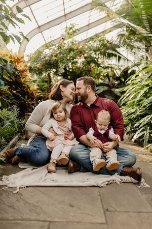 Family sits together on floor of greenhouse surrounded by lush tropical plants and greenery in Wheaton Maryland