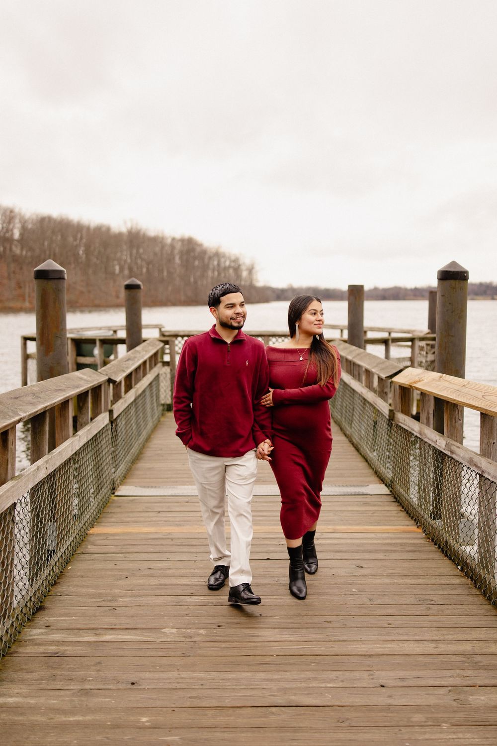 A couple in matching red sweaters walks together on a wooden pier overlooking a misty lake on a winter day in Boyds Maryland