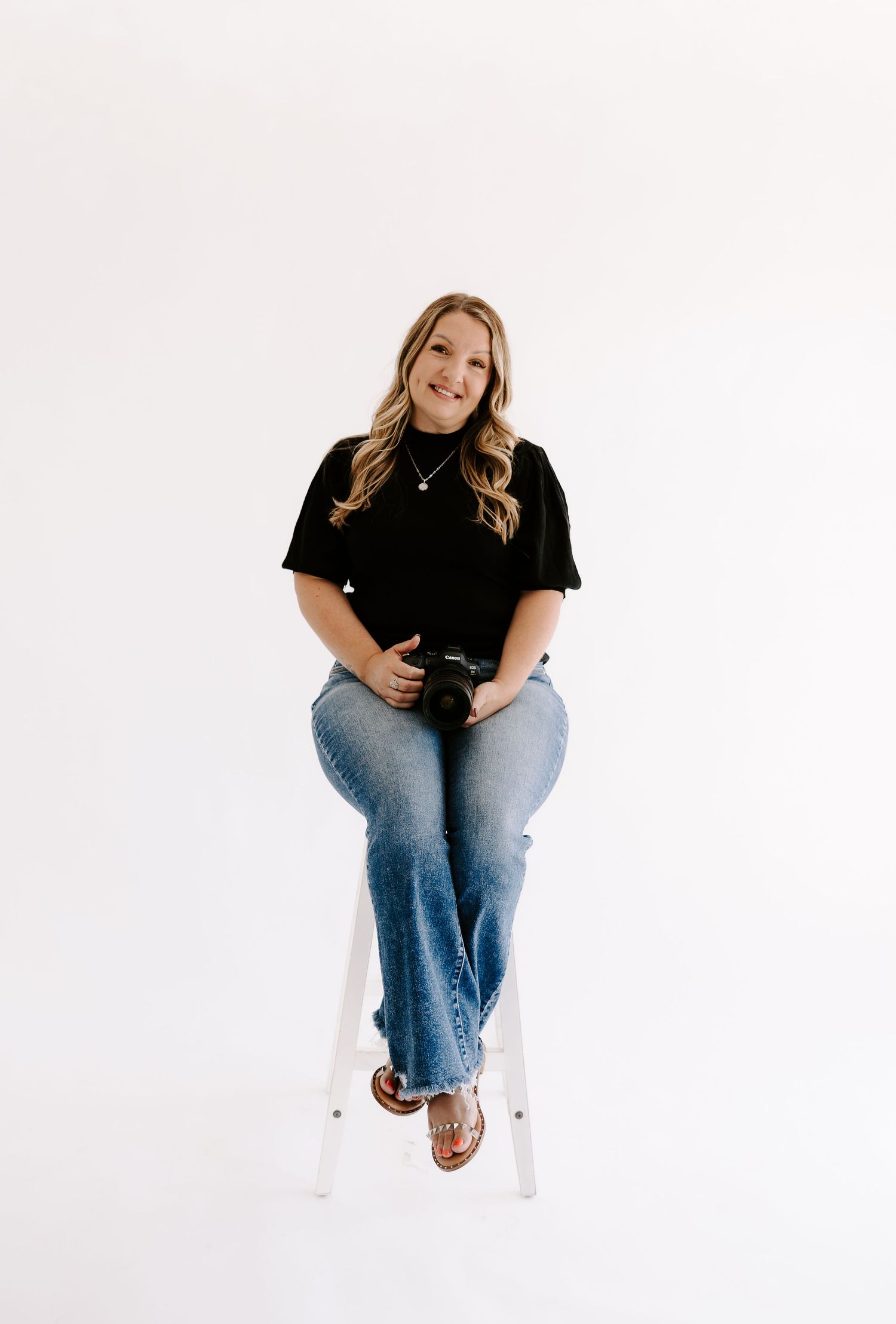 Professional wedding photographer smiling in black top and jeans holding camera on white background.