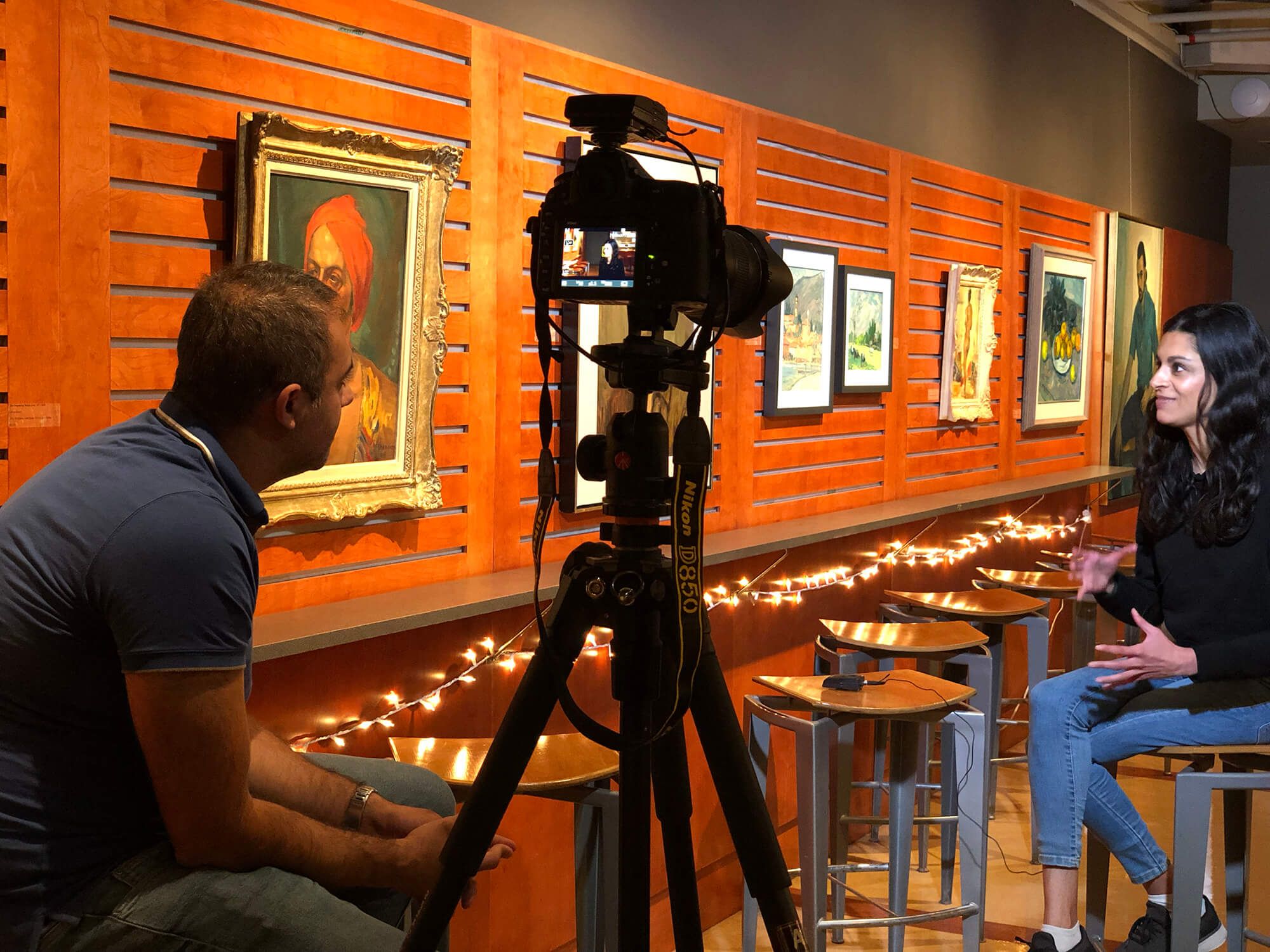 A photographer sets up camera equipment in an art gallery with orange walls displaying framed artwork. Behind the Scenes