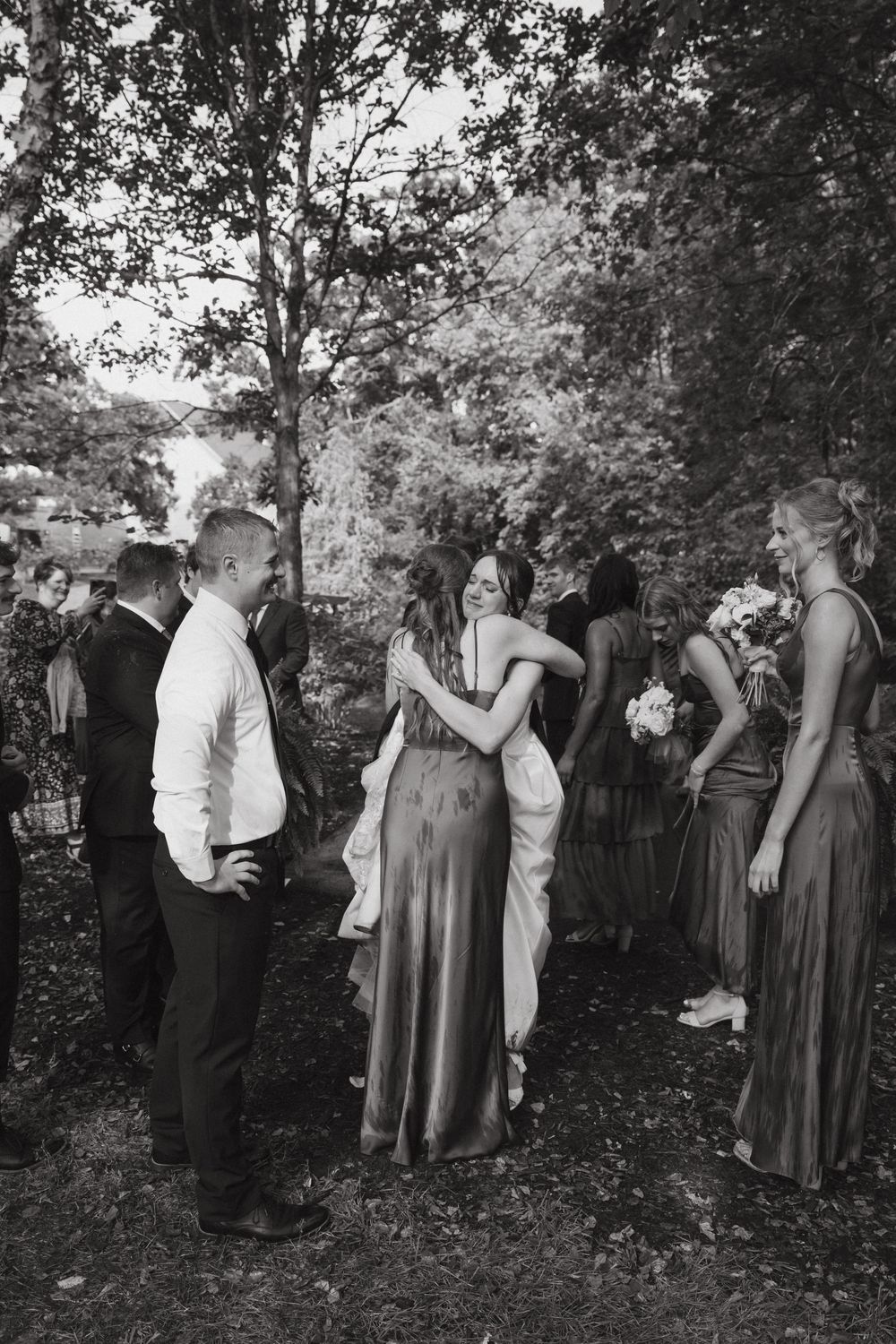 A black and white wedding photo showing a bridal party gathered outdoors under trees during a celebration.