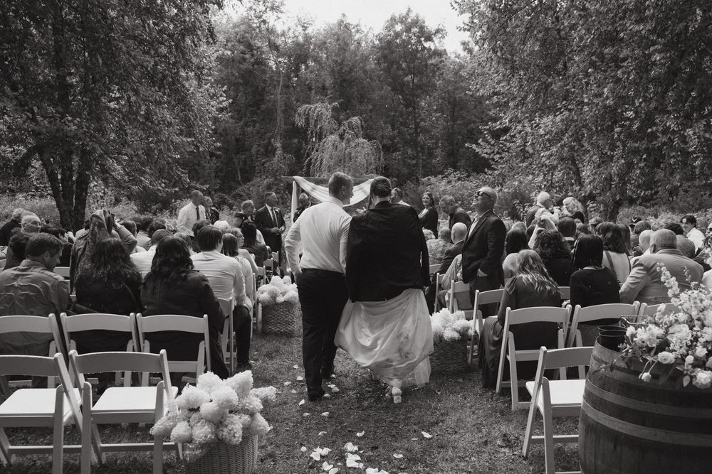 Black and white panoramic view of an outdoor wedding ceremony with guests seated in white chairs surrounded by trees.