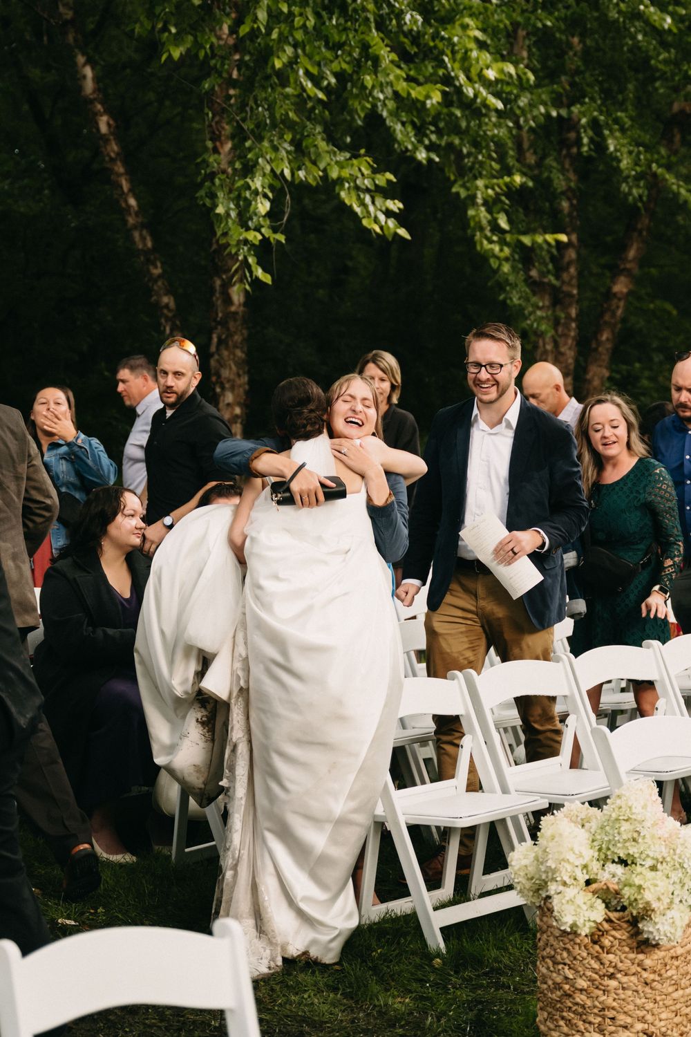 Wedding guests share emotional reactions during an outdoor ceremony with white chairs and natural greenery.