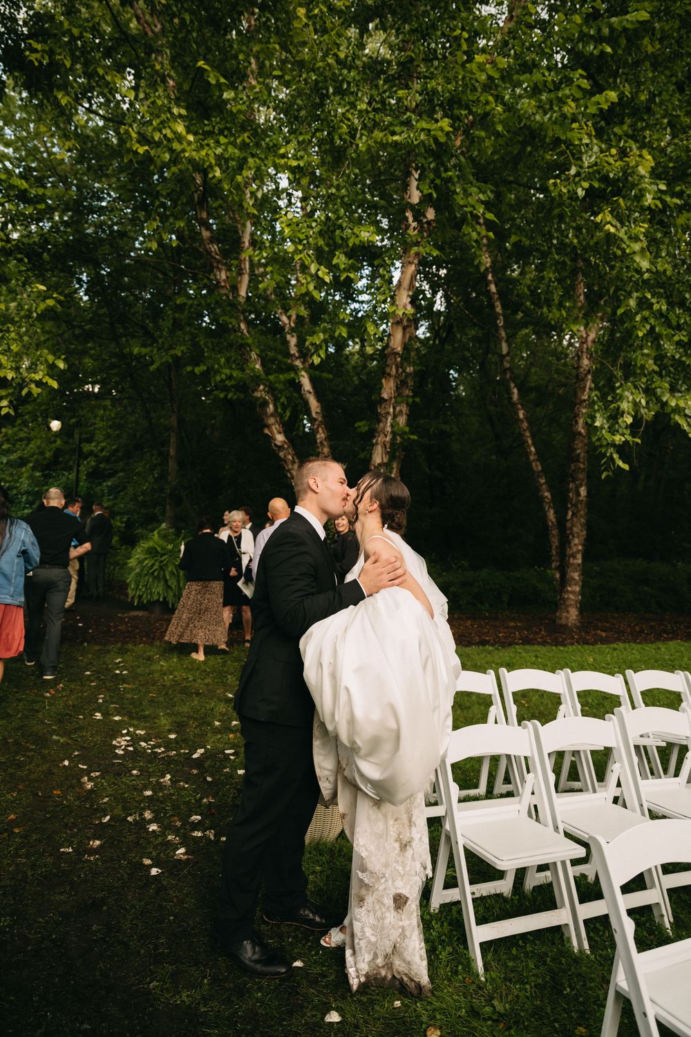 A wedding ceremony setup outdoors with white chairs arranged in rows beneath tall trees in a wooded setting.