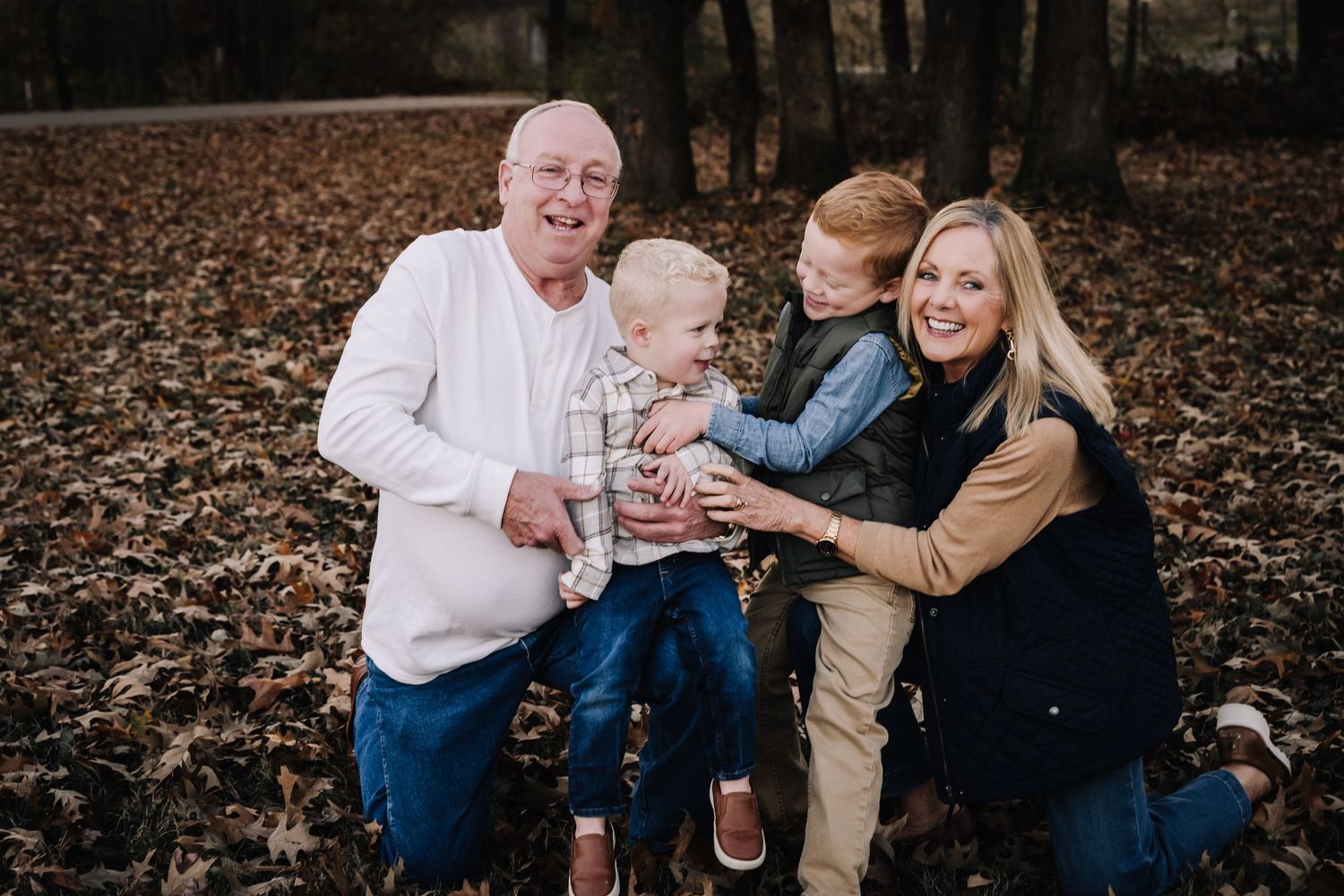 Grandparents and grandkids play together outdoors in a wooded area covered with fallen autumn leaves. Family photographer in Middle Tennesse