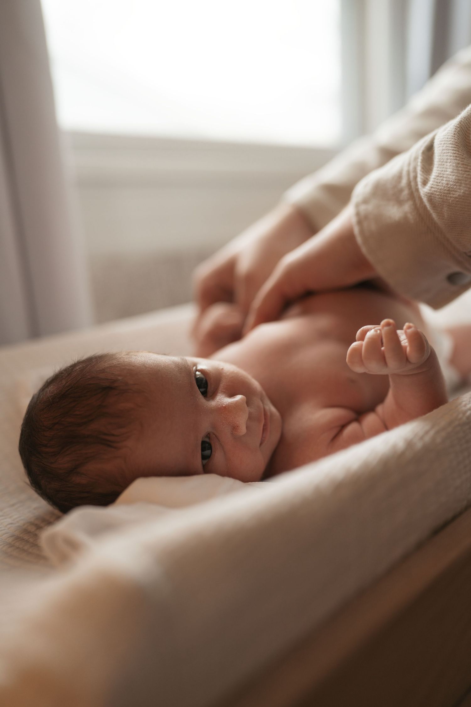 A newborn baby rests on soft white bedding in natural window light, creating a serene and intimate portrait during an in home photo session