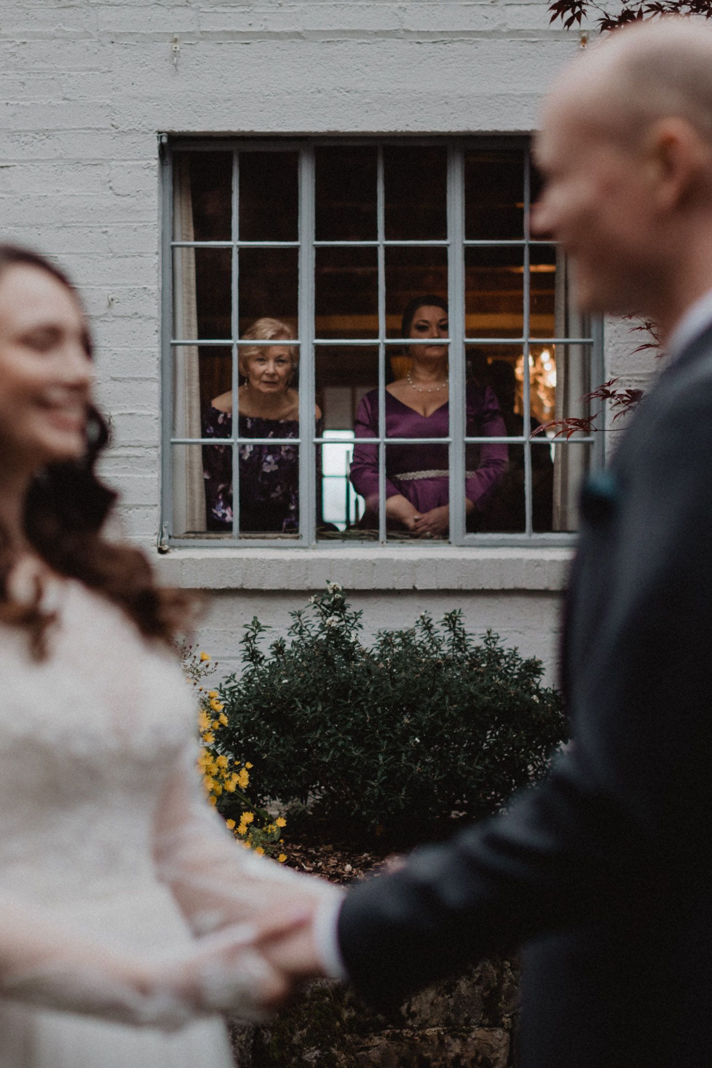Wedding guests peek through a window while watching a couple holding hands during an intimate outdoor ceremony.